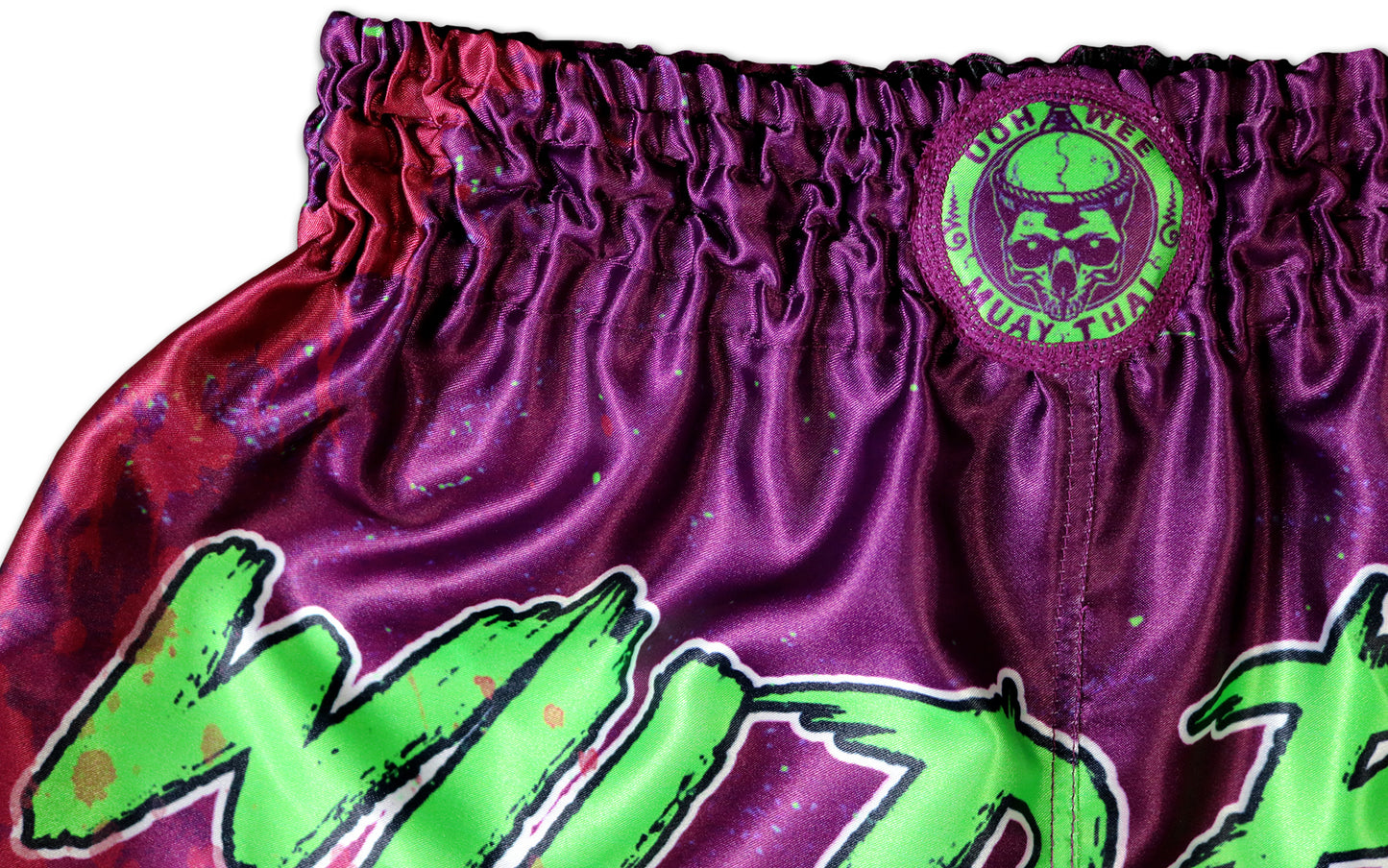 Close-up of the waistband on Wild Boy Muay Thai Shorts showing elastic purple satin fabric, neon green Oowee Muay Thai skull logo patch, and part of neon green graffiti lettering, handmade in Thailand, extreme limited edition, customizable with name.