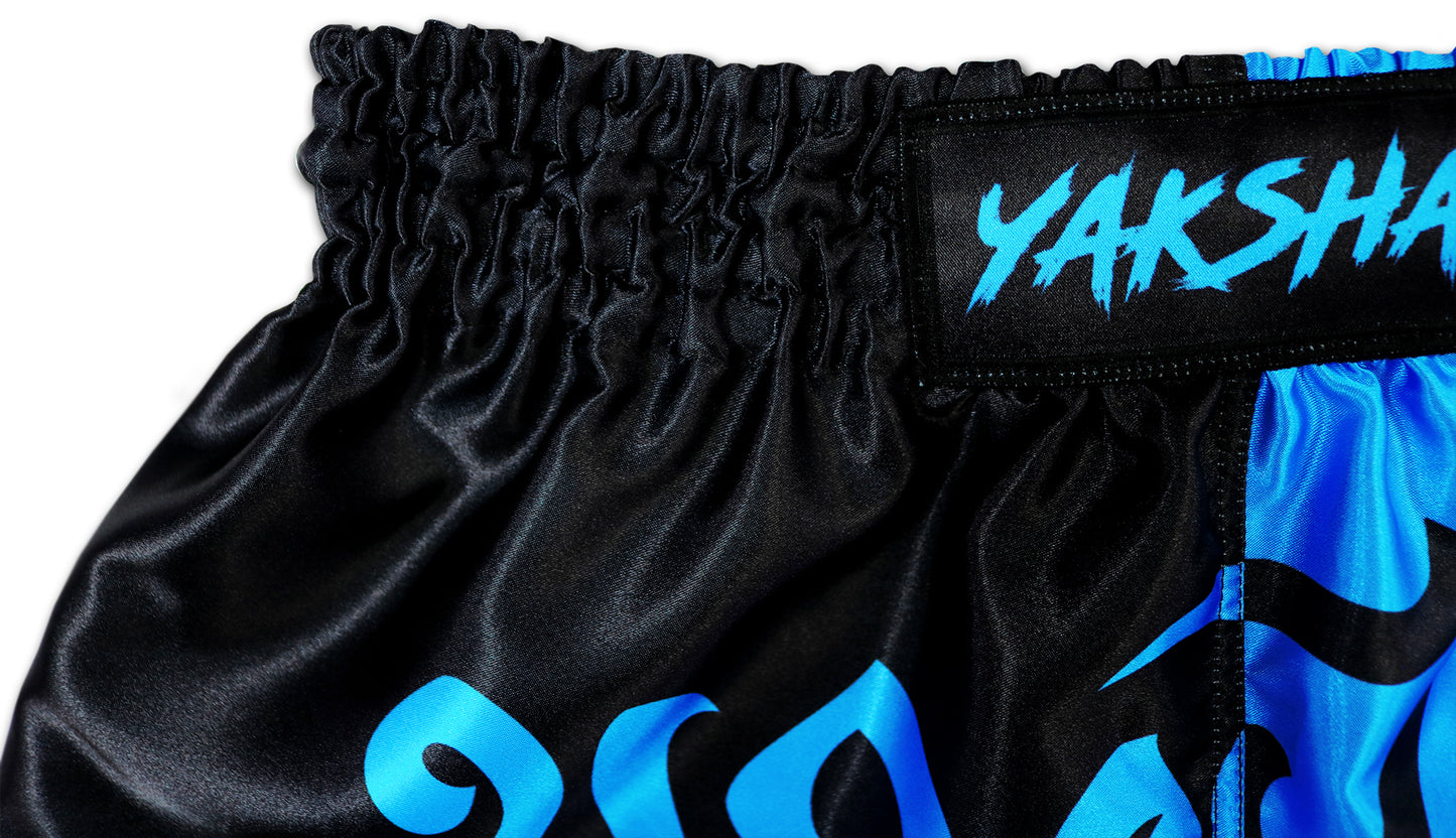 Close-up of black and blue two tone Muay Thai shorts waistband showing YAKSHA logo patch and elastic stitching detail on premium satin fabric.