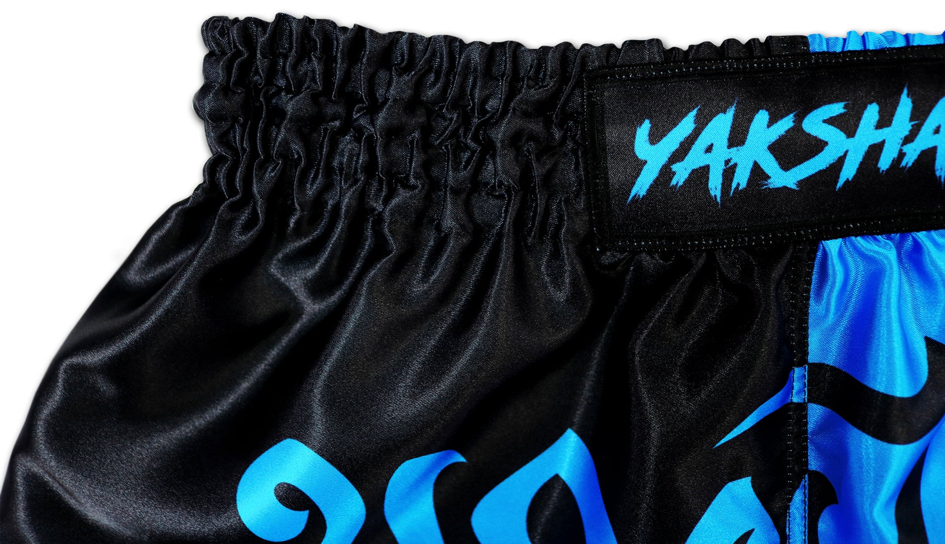 Close-up of black and blue two tone Muay Thai shorts waistband showing YAKSHA logo patch and elastic stitching detail on premium satin fabric.
