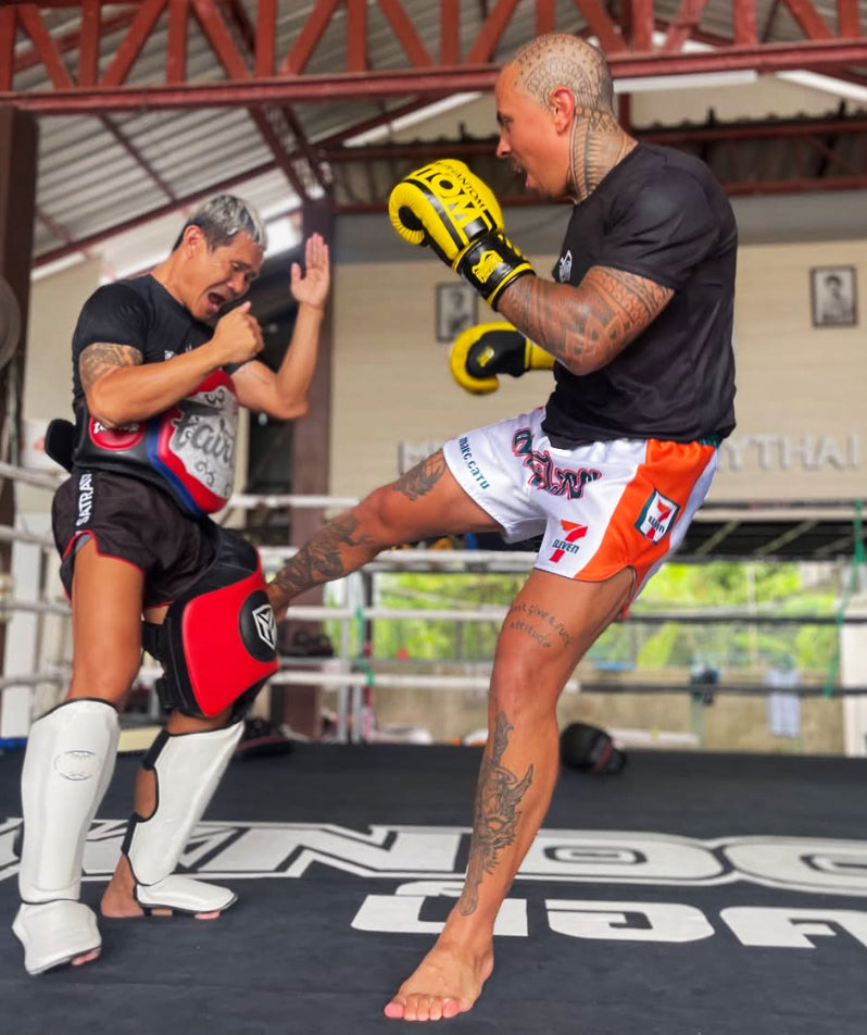 Muay Thai fighter Marc CaTu wearing  7-Eleven Muay Thai shorts delivers a kick during training in a boxing ring, with coach holding pads.