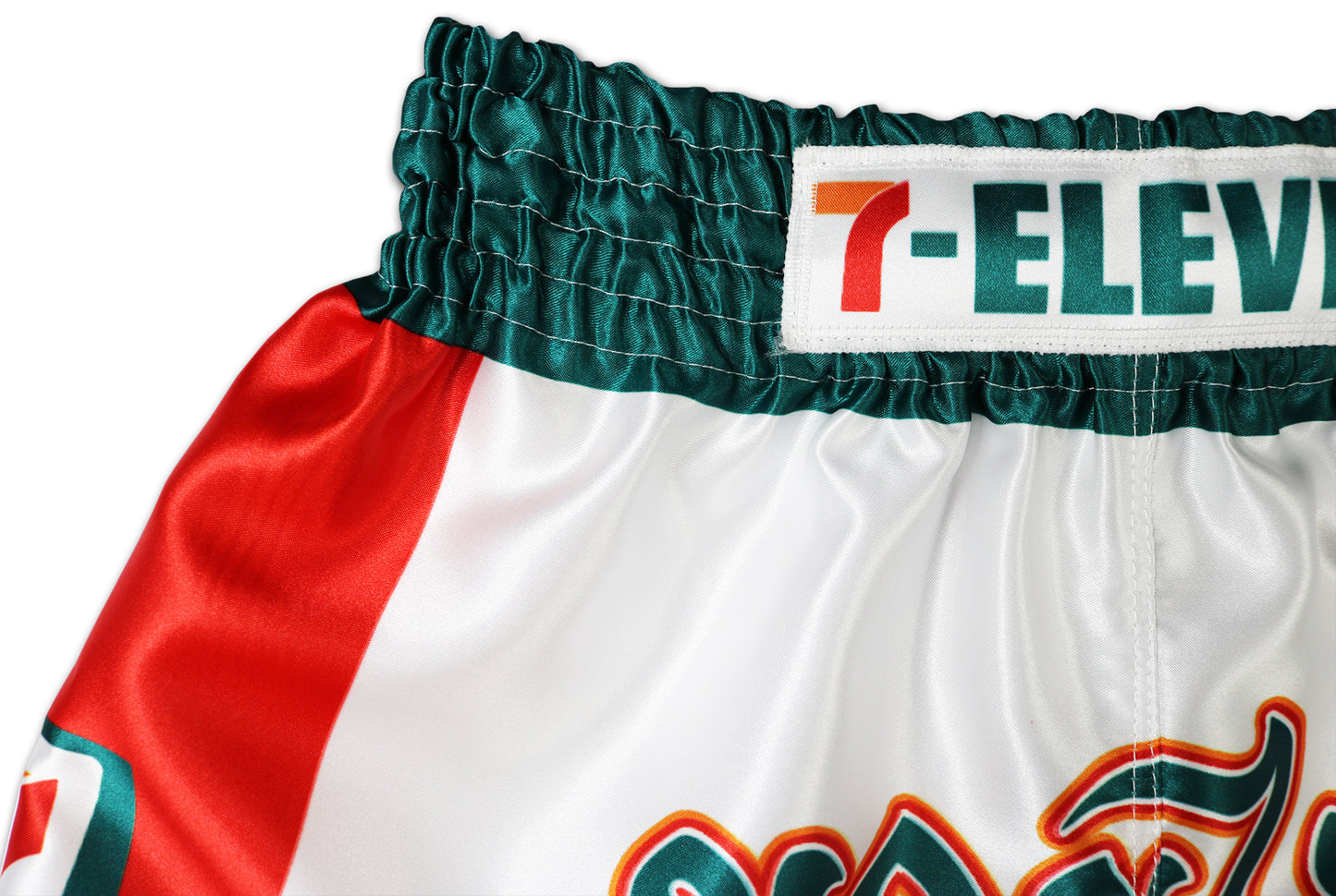 Close-up of the waistband and upper front of white Muay Thai shorts, showing green elastic band, 7-Eleven logo patch, red side panel, and part of Thai lettering.