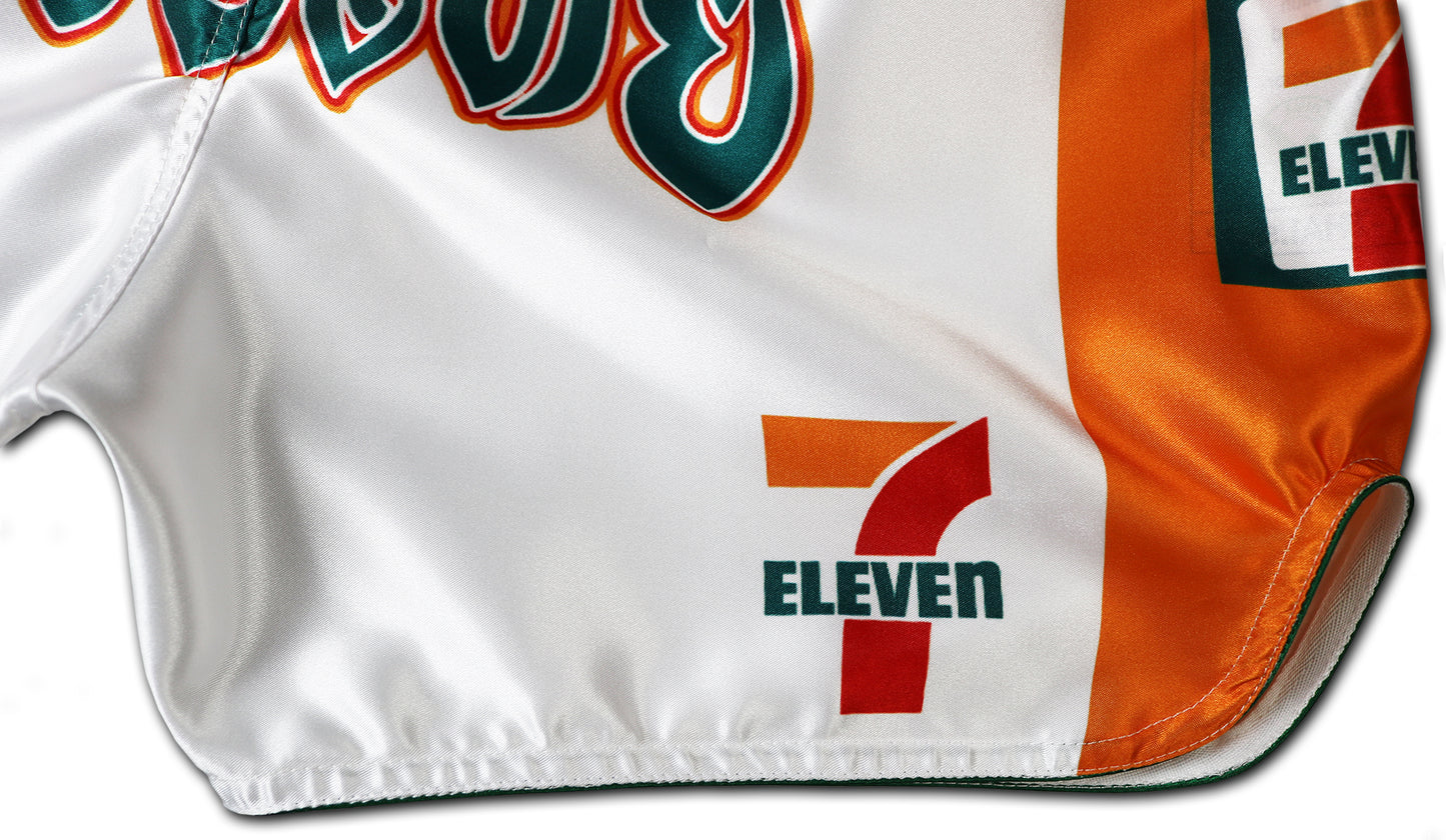 Detail picture of white Muay Thai shorts showing orange side panel, 7-Eleven logos, and part of bold Thai lettering on shiny satin fabric.
