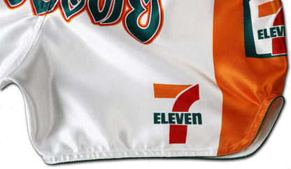 Detail picture of white Muay Thai shorts showing orange side panel, 7-Eleven logos, and part of bold Thai lettering on shiny satin fabric.