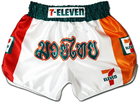 White Muay Thai shorts featuring bold green, red, and orange 7-Eleven colors, Thai lettering on the front, and 7-Eleven logos on waistband and sides.