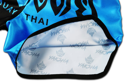 Inside view of two tone Muay Thai shorts in black and blue showing white inner lining with repeating YAKSHA logo print, handcrafted in Thailand.