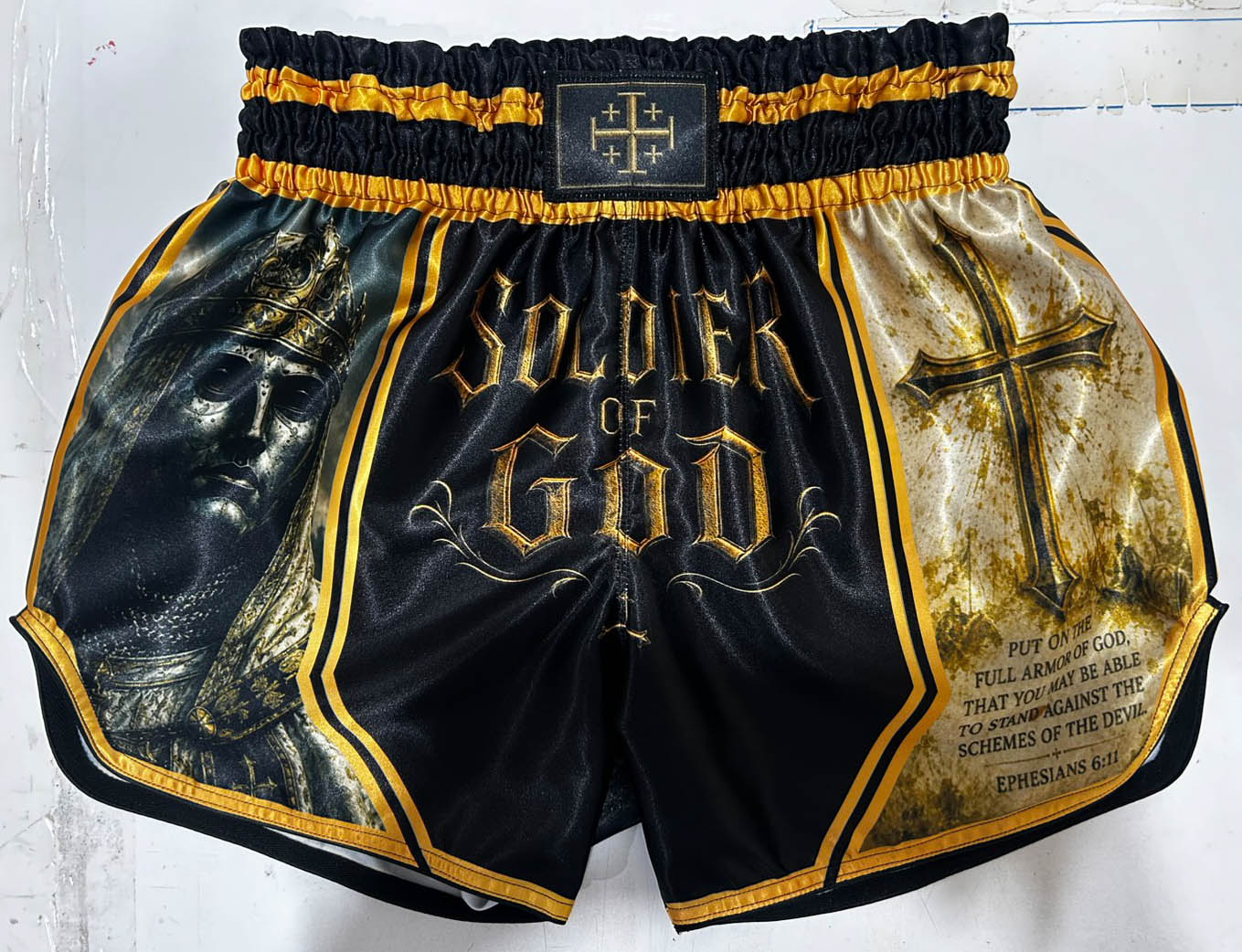 front side of a custom made muay thai shorts showing a golden soldier of god font with a krowned knight and a cross