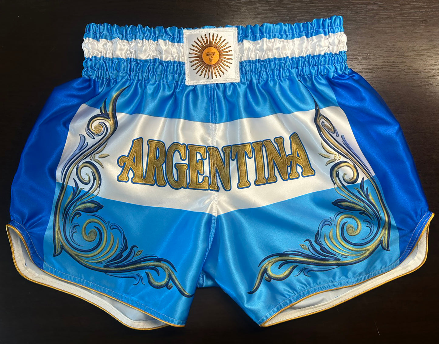 Custom-made Muay Thai Shorts Argentina Flag Design in Blue and White with Gold "Argentina" Lettering, Sun of May Patch and Ornamental Scroll Print