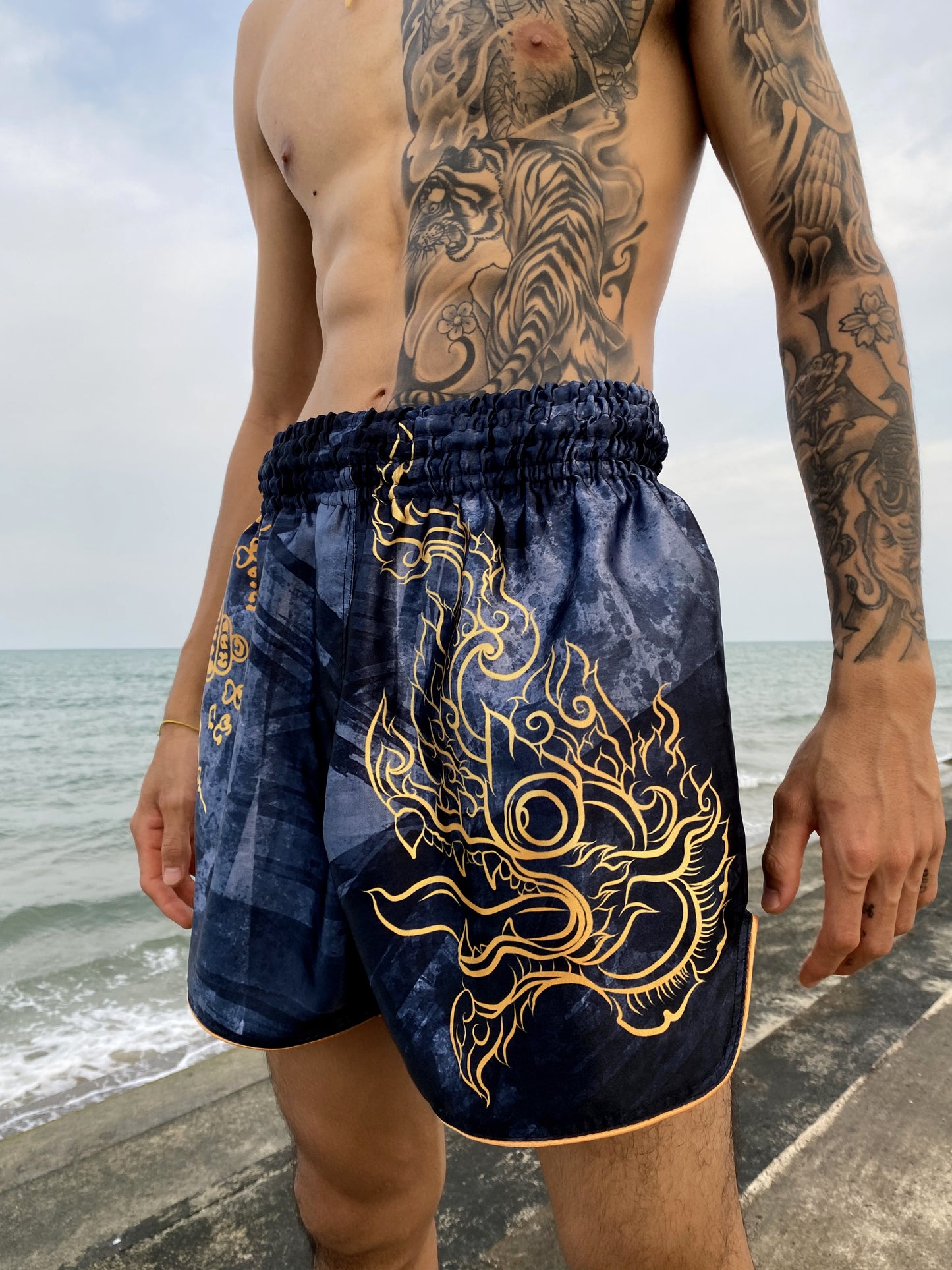 Fighter wearing Phaya Naga Muay Thai fight shorts in black at the beach, gold Naga head with flames printed on the side