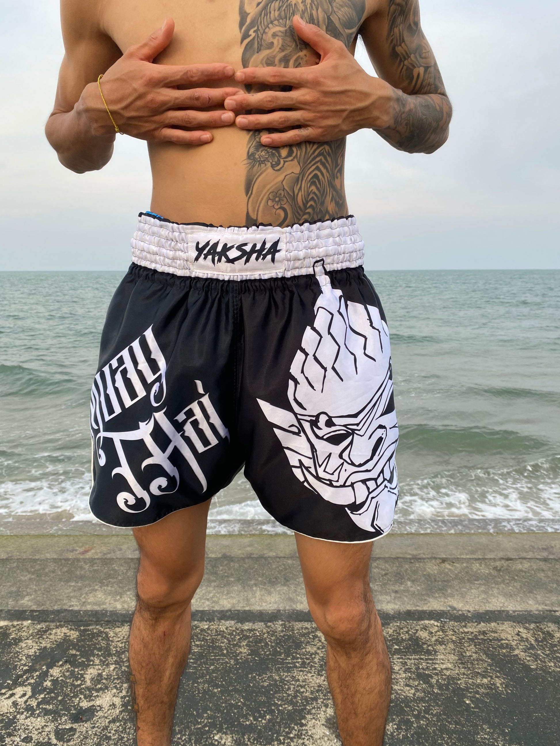 Fighter wearing YAKSHA Yin Yang black satin fightshorts with white Yaksha demon design, standing by the ocean