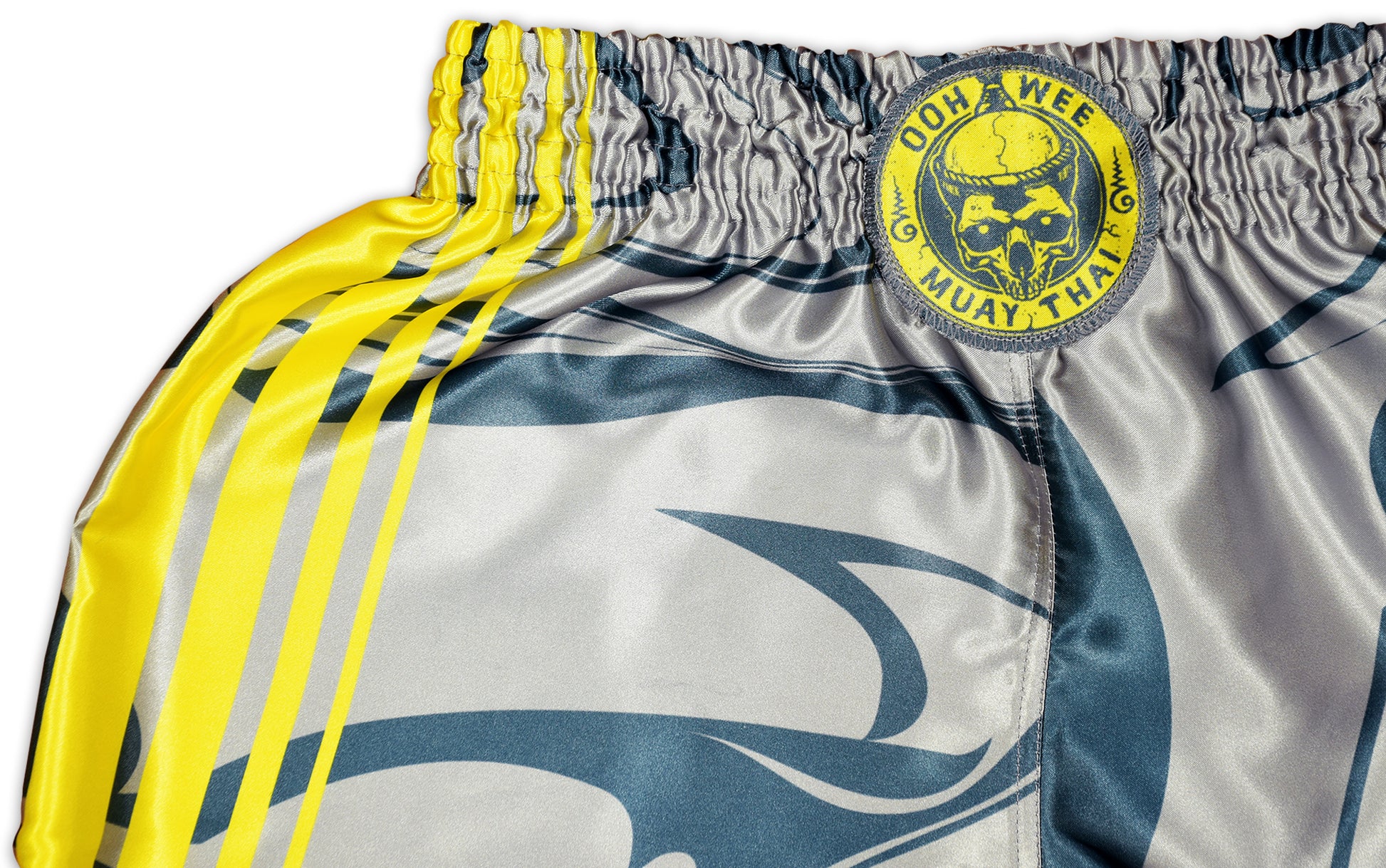 OOWEE Muay Thai Shorts detail showing the iconic OOWEE skull logo and yellow stripes on gray satin