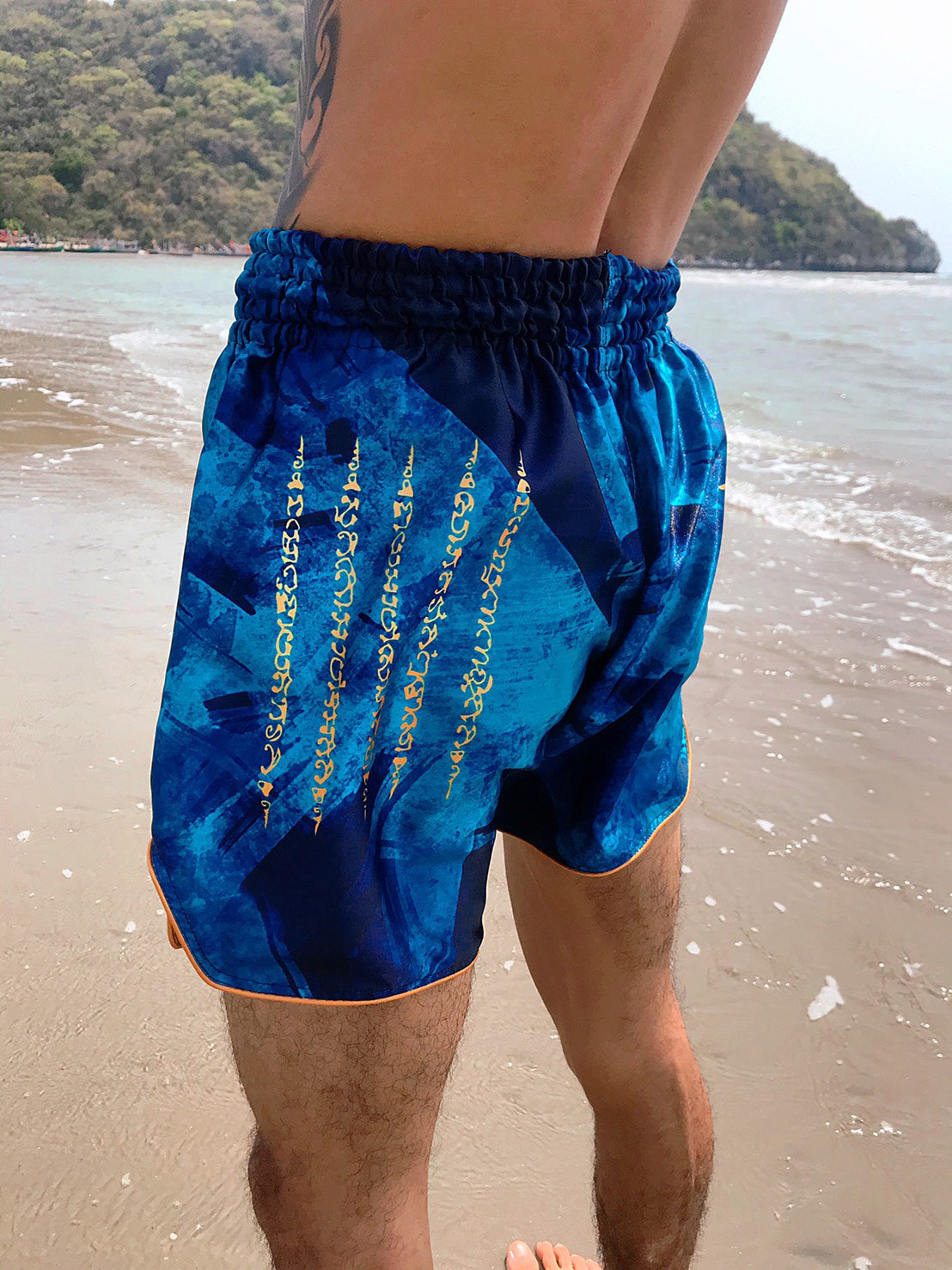 Back view of Thai boxer wearing Phaya Naga aqua blue Muay Thai Fight Shorts with gold tiger claw Sak Yant by the ocean in Thailand