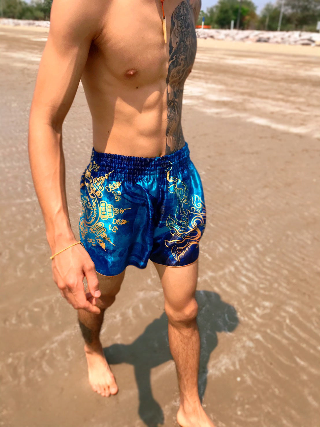 Close up of Thai fighter wearing Phaya Naga aqua blue Muay Thai Fight Shorts with gold Naga and Sak Yant design at the beach in Thailand
