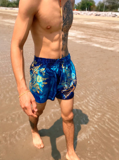 Close up of Thai fighter wearing Phaya Naga aqua blue Muay Thai Fight Shorts with gold Naga and Sak Yant design at the beach in Thailand